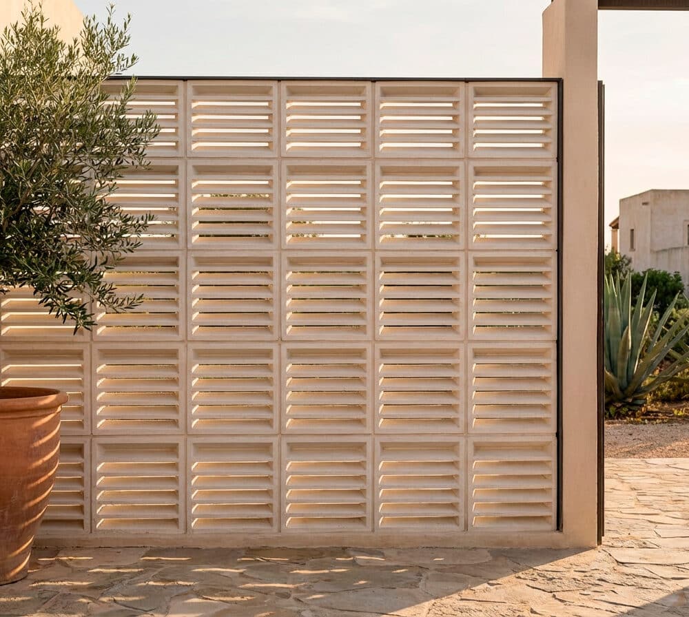 Celosía decorativa de bloques con lamas horizontales en un patio exterior. Una persiana grande junto a un olivo en maceta.