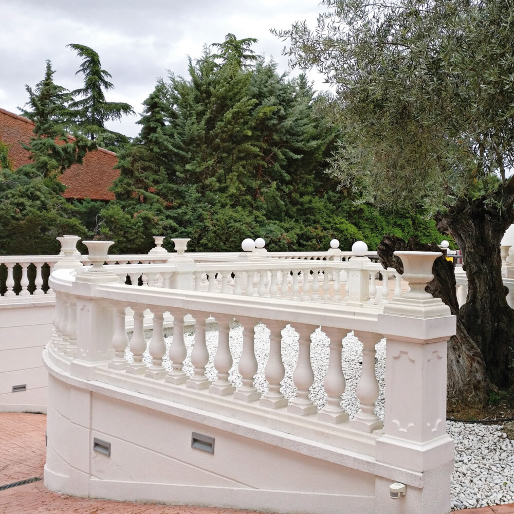 Balaustrada curva de hormigón blanco en un jardín con árboles y elementos decorativos
