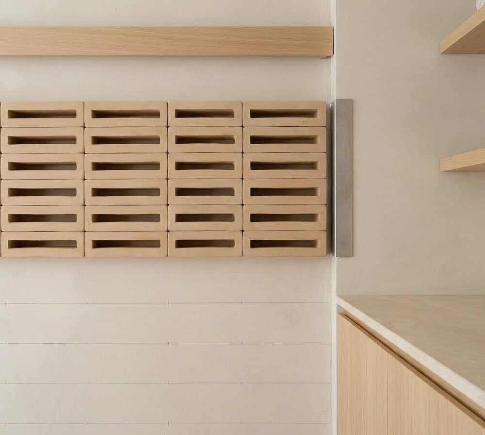 Una celosía de bloques beige con ranuras horizontales decora una pared. Estantes de madera y encimera clara a la derecha.
